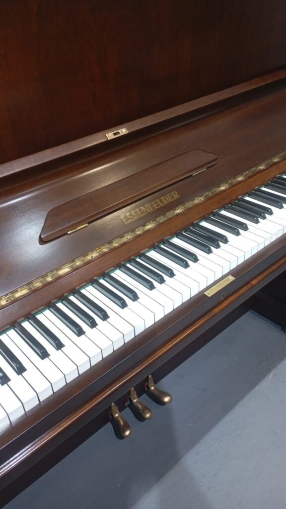 Brown upright piano with the keyboard exposed, three brass pedals below, in a room; brand plate reads ESSENFELDER/FENDER-style name on the fallboard.