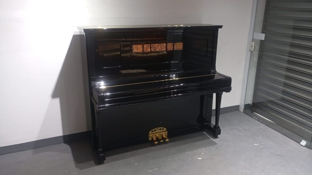 Shiny black upright piano with gold pedals placed in a concrete room by a metal roll-up door