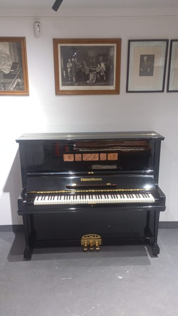 Black upright piano with gold pedals in a gallery room, framed black-and-white art on the wall behind it.